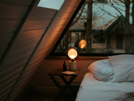 A bed under a sloping wooden roof lit by a bedside lamp