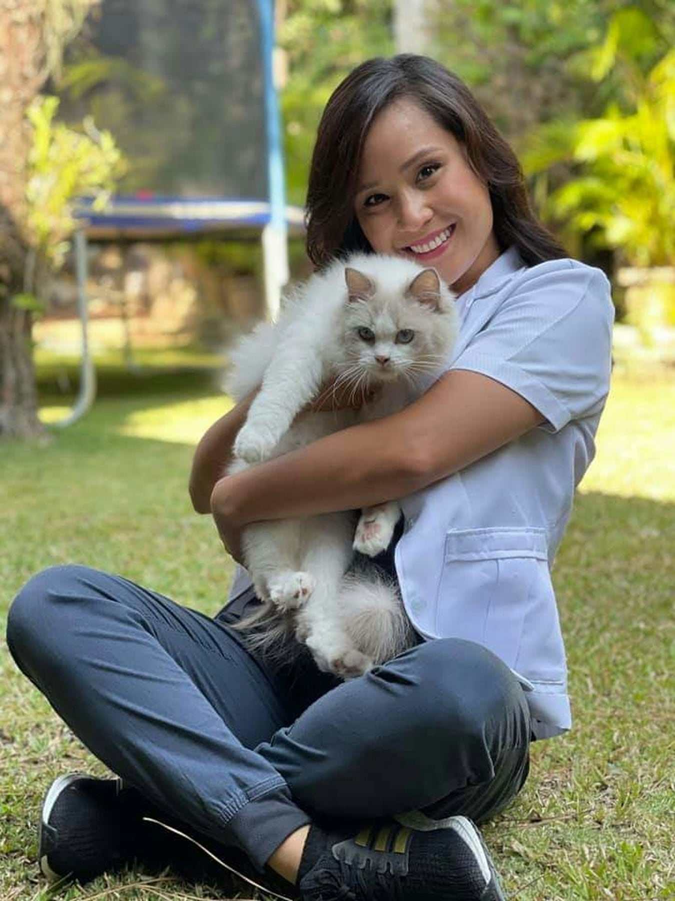 A smiling vet in a blue uniform sitting on the grass cuddling a fluffy white cat