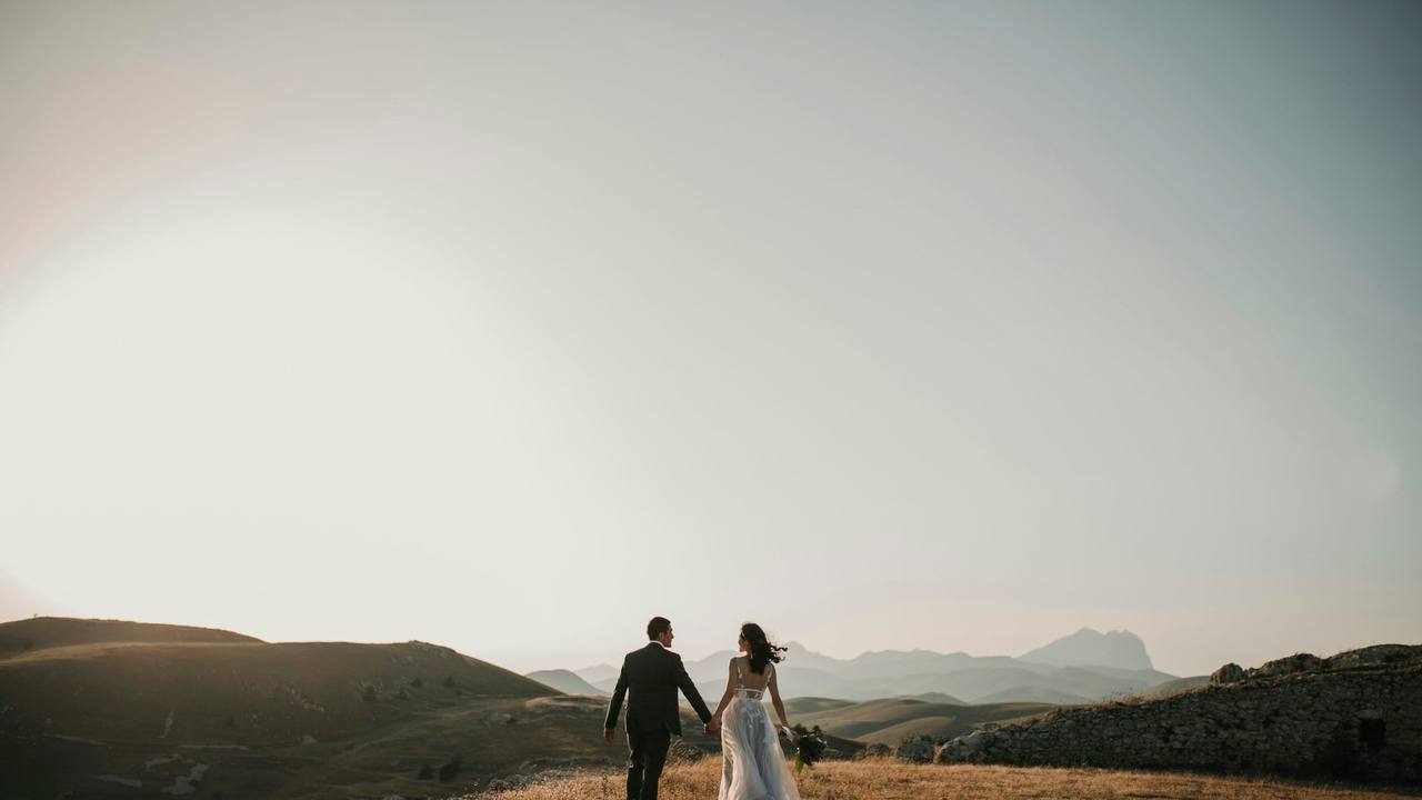 A bride and groom walking hand in hand across a wide open hillside