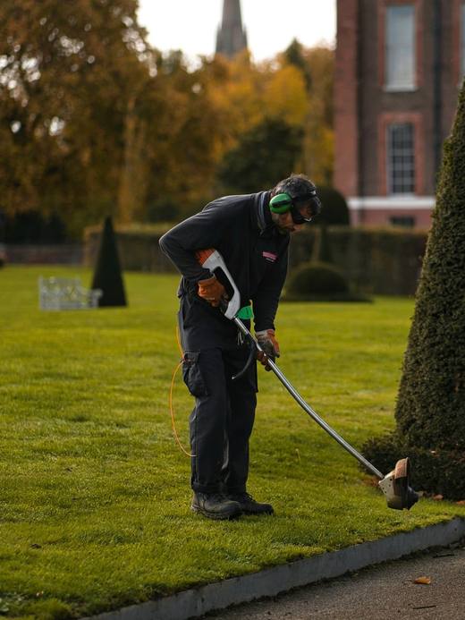 A gardener trimming a cone shaped topiary on a lawn